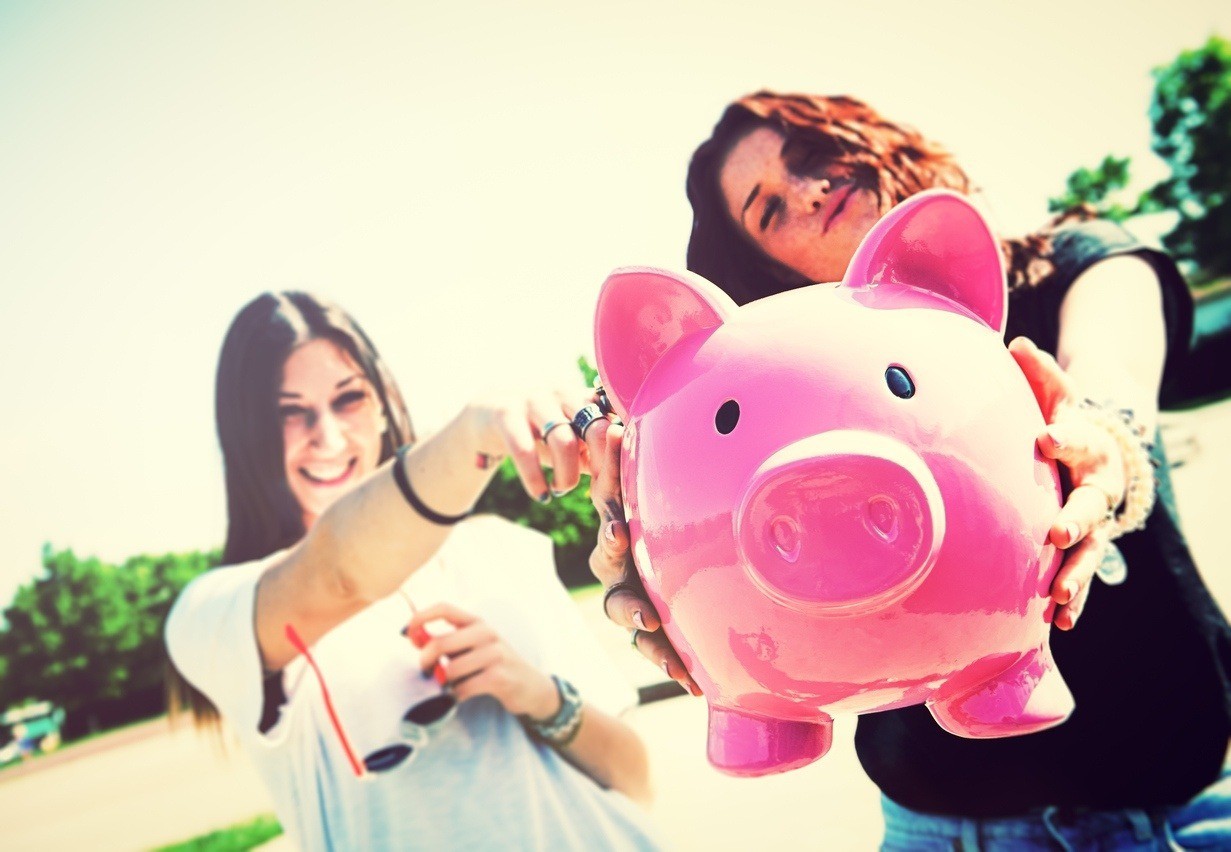 Two young funny woman holding piggy bank in hands, selective focus, outdoors