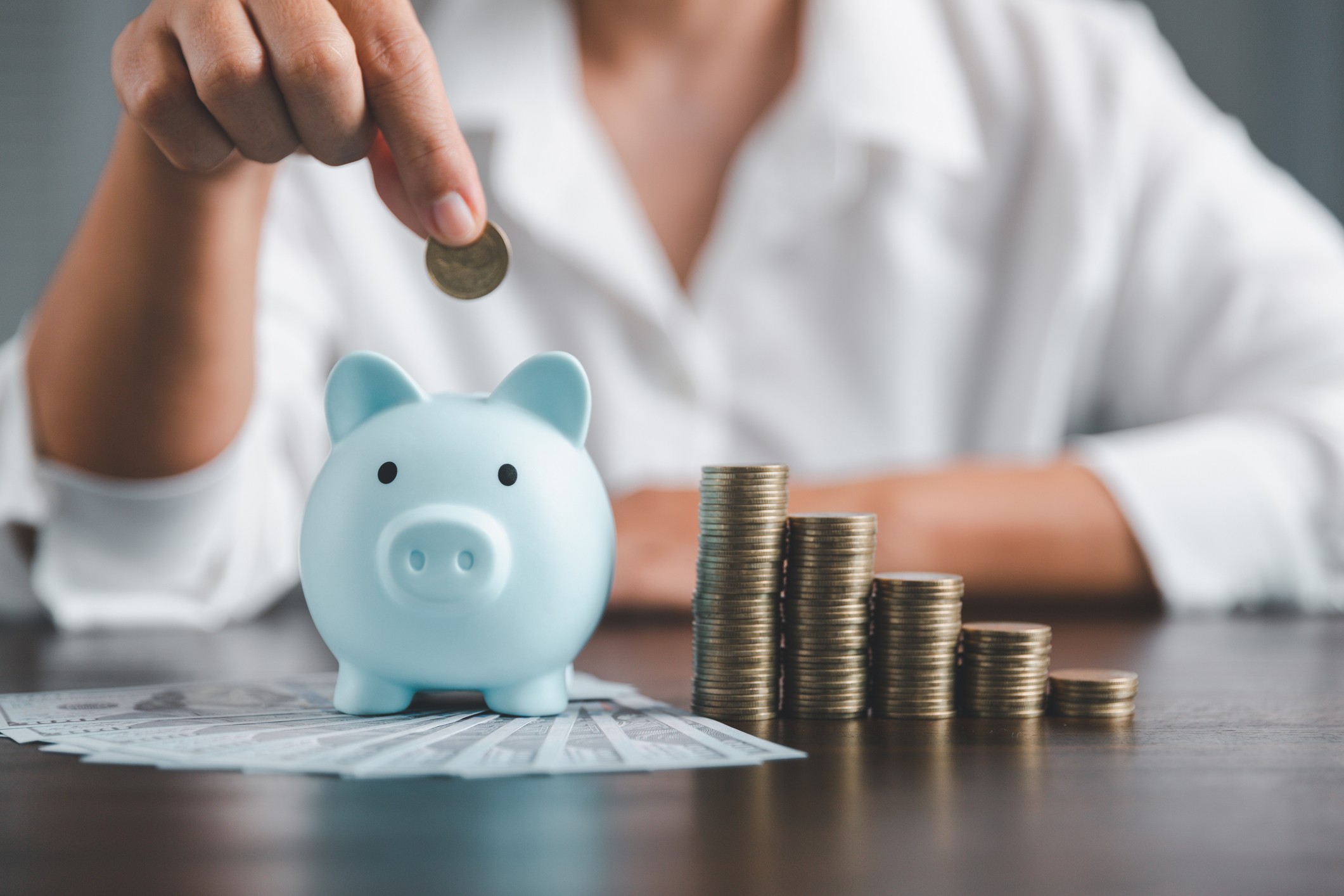 A woman depositing a coin into a blue piggy bank while sitting at a table with coins and money