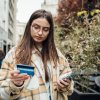 A college student holding her credit card and using a smartphone while walking in the city