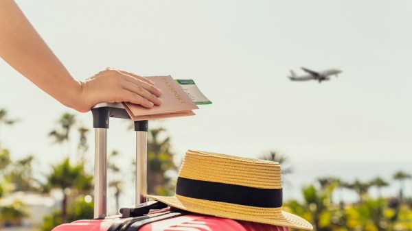 Woman with pink suitcase and passport with boarding pass standing on passengers ladder of airplane opposite sea with palm trees.