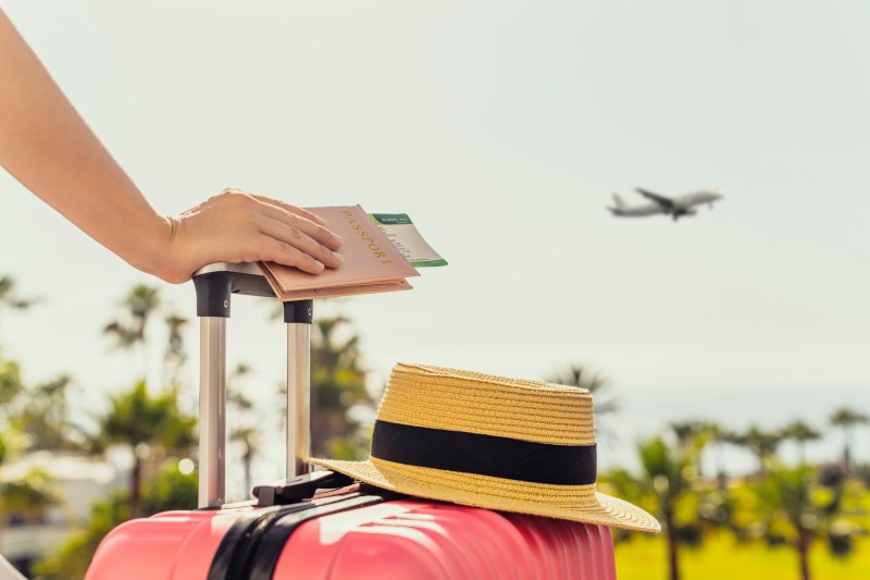 Woman with pink suitcase and passport with boarding pass standing on passengers ladder of airplane opposite sea with palm trees.