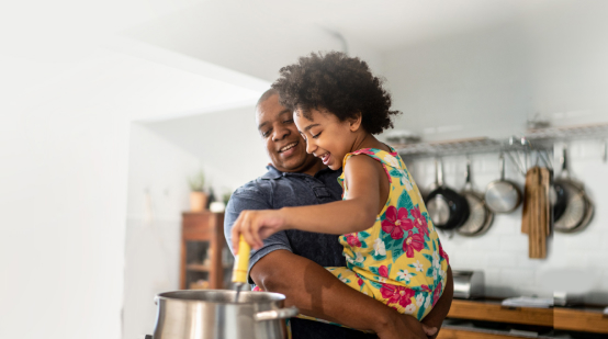 smiling dad holding smiling child while they stir a pot in a kitchen.