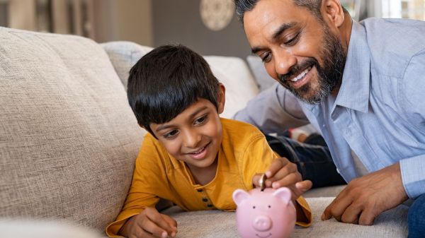 Smiling kid adding coin in piggybank while lying on couch with dad at home