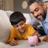 Smiling kid adding coin in piggybank while lying on couch with dad at home