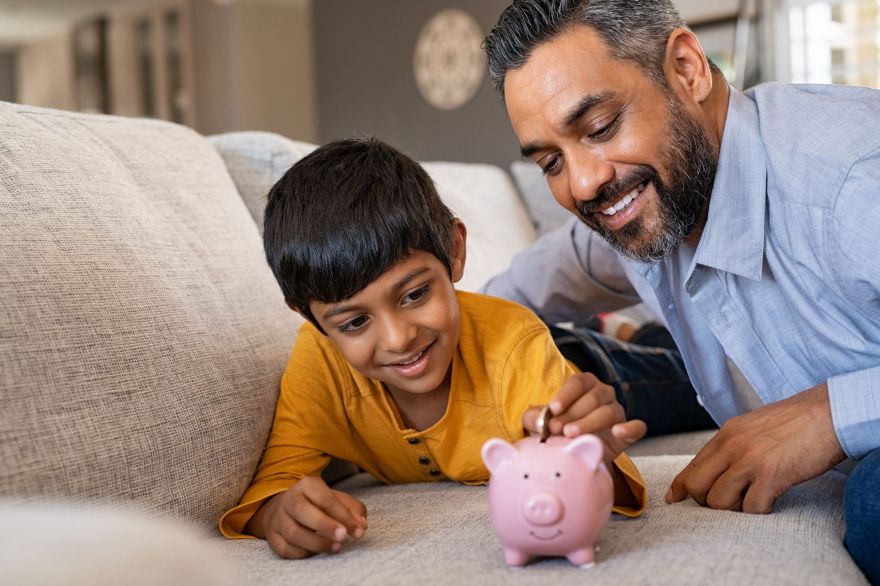 Smiling kid adding coin in piggybank while lying on couch with dad at home