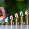 A hand holds a home model to put on a stack coin with other house models with coin stacks