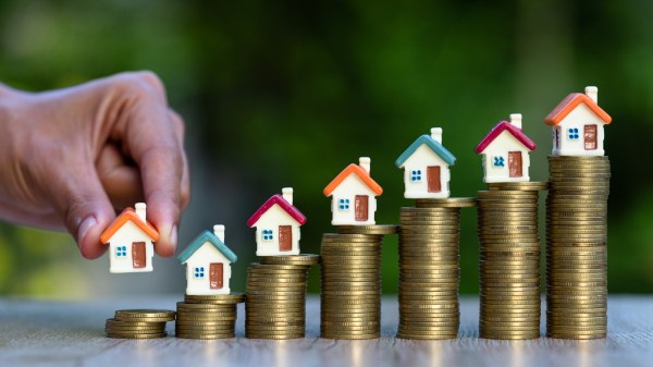 A hand holds a home model to put on a stack coin with other house models with coin stacks